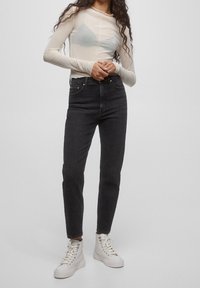 Jeans negros de talle alto combinados con una blusa transparente de manga larga. La modelo lleva zapatillas blancas de caña alta. Características incluyen un diseño ajustado y costuras limpias.