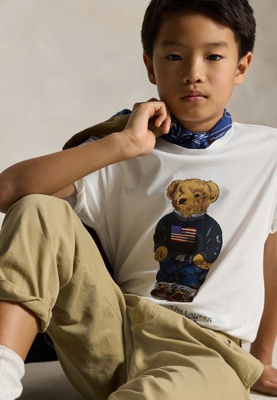 Young boy wearing beige pants, white t-shirt with teddy bear print holding an American flag sweater, and a blue patterned neck scarf.