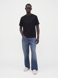 Άνδρας στέκεται φορώντας μαύρο γραφικό t-shirt, ξεθωριασμένο μπλε φαρδύ τζιν παντελόνι και λευκά αθλητικά παπούτσια σε απλό φωτεινό φόντο.