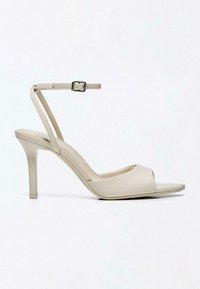 Sandale à talon haut beige avec bout ouvert, talon aigu fin et bride de cheville avec boucle argentée sur fond blanc.