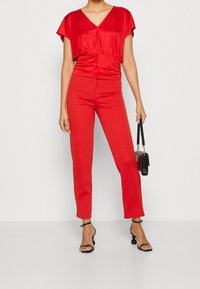 Rød satin-top med V-hals og korte ærmer, kombineret med røde, tætsiddende bukser. Tilbehør inkluderer en sort håndtaske og sorte hæle med stropper.