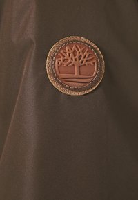 Tissu marron à texture lisse, doté d'un patch circulaire en cuir embossé d'un motif d'arbre, bordé d'un bord en cuir marron clair texturé.