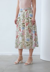 Floral midi skirt in cream, featuring various colored flowers, green leaves, and an elastic waistband. Paired with white strappy block-heel sandals.