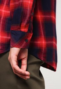 Rood en marineblauwe geruite flanellen overhemd met een knoopmanchet. De stof heeft een zachte textuur en een duidelijk ruitpatroon. Subtiele afgeronde zoom.