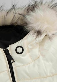 Kremowa pikowana kurtka z czarnym zamkiem błyskawicznym i odpinanym kapturem, wyposażona w jasnobeżowe futro syntetyczne z ciemnymi końcówkami.