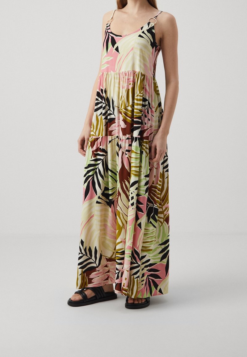 Femme portant une robe maxi sans manches imprimée de feuilles tropicales roses, vertes et noires, avec des bretelles à anneaux et des sandales plates noires.