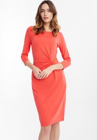 Korallefarbenes Kleid mit drei Viertelärmel, das eine Drehung in der Taille und eine glatte Textur aufweist. Einfaches, knielanges Design.