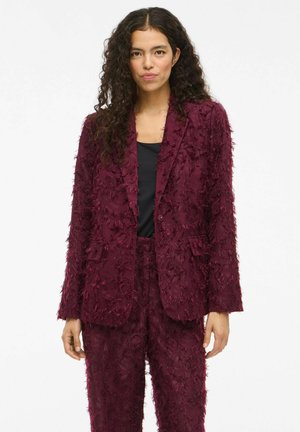Bordeaux gestructureerd pak met rafelige stofdetails; getailleerde blazer met notch-uitsnijdingen en bijpassende broek, gecombineerd met een zwarte top.