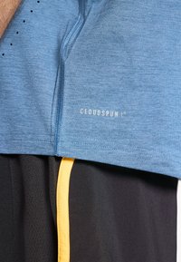 Lyseblå atletisk skjorte med en glat tekstur, der har en lynlåsåbning i halsen og "CLOUDSPUN" trykt i hvid. Kombineret med sorte shorts og gule detaljer.