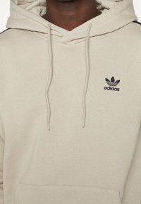 Beige Adidas-hoodie med dragsko, känguruficka och liten svart Adidas-logotyp på vänster bröst, buren av en person.