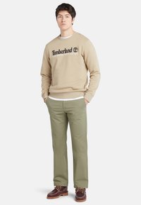 Joven con sudadera beige de Timberland, pantalones verdes oliva y zapatos náuticos de cuero marrón, de pie con las manos en los bolsillos.