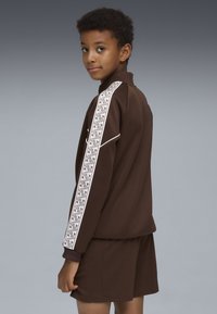 Chándal atlético marrón, que incluye una chaqueta con cremallera y panel lateral blanco con patrón, y pantalones cortos marrones. Tejido texturizado, puños acanalados.