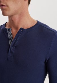 Marineblå langærmet henley trøje med en knaplukning og et stribet stofakcent samt en rund halsudskæring. Glat tekstur.