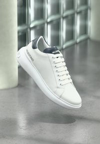 Λευκό αθλητικό παπούτσι Valentino με μαύρη ταμπέλα στο πίσω μέρος και λογότυπο στο πλάι και στη γλώσσα, εμφανίζεται να αιωρείται πάνω από μια αντανακλαστική τσιμεντένια επιφάνεια.