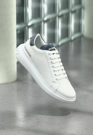 Biały sneaker Valentino z czarną wstawką na pięcie oraz logo po boku i na języku, unoszący się nad refleksyjną betonową podłogą.