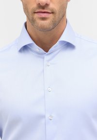 Camicia di dress azzurro chiaro con un motivo texturizzato, apertura frontale con bottoni e colletto a punta. Caratteristiche includono bottoni bianchi e una vestibilità su misura.