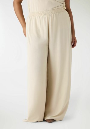 Beżowe spodnie o szerokich nogawkach z elastycznym pasem, noszone z brązowymi butami z noskiem w kształcie ostro zakończonego szpicu, na osobie z jedną ręką w kieszeni, a drugą wzdłuż ciała.