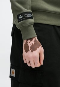 Φούτερ ελιάς με ριμπ μανσέτες και μαύρη ετικέτα στο αριστερό μανίκι. Το χέρι έχει καφέ απόχρωση δέρματος· μαύρο παντελόνι φαίνεται από κάτω.