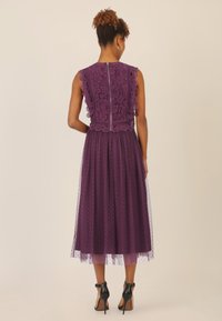 Abito in pizzo viola con corpino aderente, bordi scallop e gonna in tulle con pois, completato da scarpe con tacco nere.