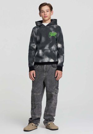 Ragazzo adolescenti che indossa un hoodie nero e bianco con motivo tie-dye e una grafica di un controller da gioco verde, pantaloni cargo grigi e scarpe beige senza lacci.