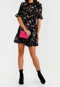 Kvinna som bär en kort svart blommig klänning, svarta ankelskor och håller en liten ljusrosa axelväska.