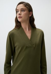 Top lungo a maniche con scollatura a V di colore verde oliva, con una texture liscia. Presenta un dettaglio di cucitura sul colletto e una vestibilità rilassata.