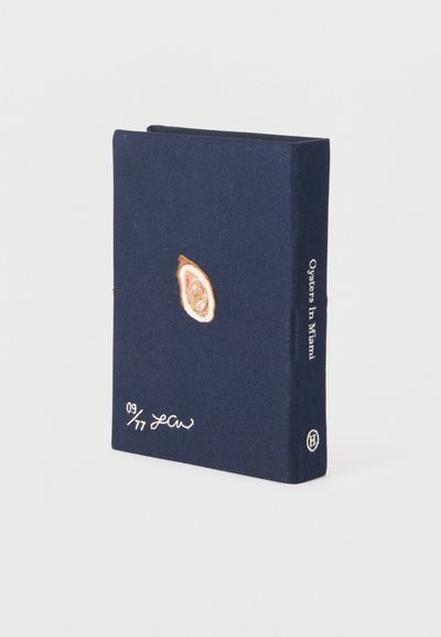 Βιβλίο σε ναυτικό μπλε χρώμα με κεντημένο στρείδι στο εξώφυλλο, τίτλο "Στρείδια στο Μαϊάμι" στην ράχη και χειρόγραφη υπογραφή με αριθμούς.