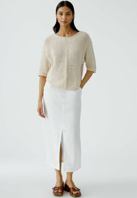 Beiger Strickpullover mit kurzen Ärmeln, kombiniert mit einem weißen Leinenrock mit einem Schlitz vorne. Modell trägt braune Plateausandalen.