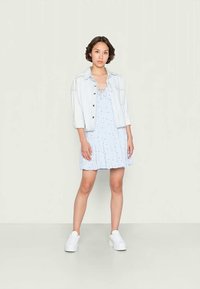 Vestido sem mangas azul claro com bolinhas brancas, bainha com babados e detalhe de laço na frente, combinado com uma jaqueta de denim clara e ténis brancos.