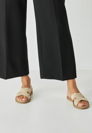 Offene Pantoletten aus beigem, geflochtenem Stoff mit einem spiralförmigen Design auf dem Riemen und einer flachen, braunen Sohle.