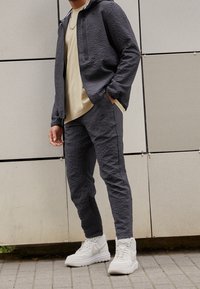 Man som bär en strukturerad mörkgrå träningsoverall och vita högskurna sneakers, står mot en kaklad vägg på ett belagt golv.