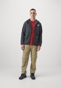 Szary rozpinany polar, czerwony t-shirt, beżowe spodnie cargo z zamkiem, oraz czarne sneakersy. Model stoi na tle w neutralnych kolorach.