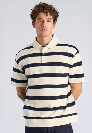 Jonge man met krullend haar, gekleed in een crèmekleurig poloshirt met dikke zwarte horizontale strepen, staand tegen een effen lichte achtergrond.