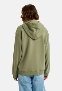 Överdimensionerad olivgrön hoodie med mjuk textur, utrustad med dragsko i huvan och ribbade ärmmuddar. Matchas med ljusblå denimjeans.