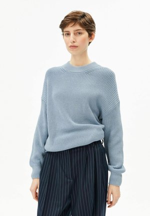 Hellblauer Strickpullover mit rundem Ausschnitt und gerippten Bündchen, kombiniert mit dunkel gestreiften Hosen. Der Stoff hat eine strukturierte Oberfläche.
