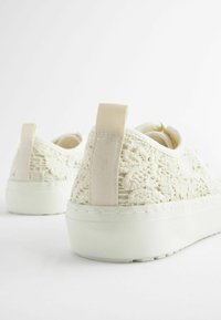 Zapatillas de encaje crema con parte superior de tejido, pestaña elástica en el talón y suela de goma. Presenta un detallado patrón intrincado y un diseño limpio y minimalista.