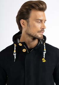 Fekete kapucnis dzseki, sárga gombokkal és mintás zsinórokkal. Simává textúrájú, finom logóval a mellkason.