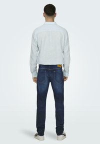 Camicia a maniche lunghe blu chiaro, jeans blu scuro con etichetta gialla e sneakers bianche. I jeans presentano dettagli di cucitura e una vestibilità slim.