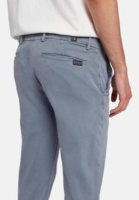 Lichtblauwe broeken van zacht materiaal, met achterzakken en een klein merkembleem op de tailleband. Soepele structuur en een pasvorm op maat.
