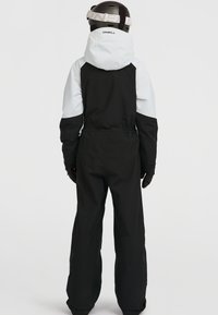 Traje de esquí en blanco y negro con mangas largas, cintura elástica y gran capucha. Acompañado de un casco marrón y guantes.