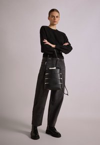 Musta nahkainen ämpärilaukku vaakasuorilla hopeisilla yksityiskohdilla, nyörisuljenta ja pitkä säädettävä hihna. Mallilla on päällä musta neule ja farkut.