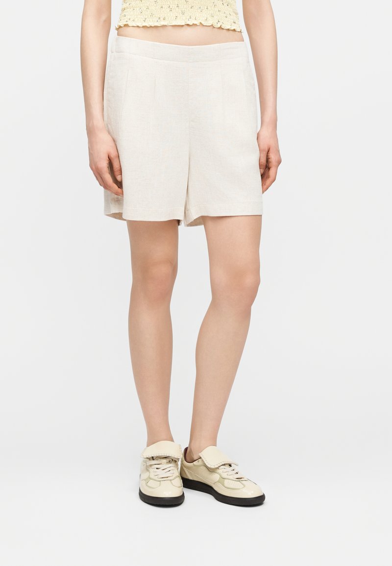 Femme portant un short crème mi-cuisse, un haut court jaune et des baskets crème à semelles noires sur fond blanc.