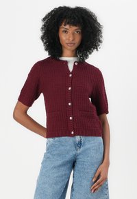 Cardigan corto a maniche corte di colore borgogna, con motivo testurizzato, chiusura a bottoni e due tasche frontali, abbinato a pantaloni in denim azzurro chiaro.