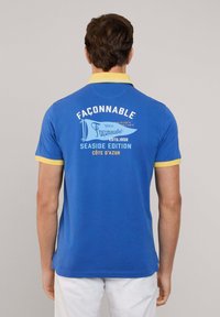 Niebieska koszulka polo z żółtym kołnierzykiem i akcentami na rękawach. Z tyłu znajduje się napis: "FAÇONNABLE", "SEASIDE EDITION" oraz dekoracyjna grafika flagi.