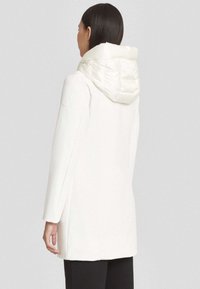 Donna con capelli scuri e lunghi che indossa un lungo cappotto bianco con un ampio cappuccio imbottito, vista di spalle su uno sfondo neutro.