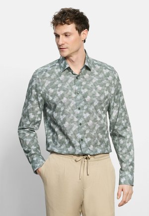 Mann trägt grünes, langärmliges Hemd mit Blumenmuster und beige Hose mit Kordelzug, steht mit einer Hand in der Tasche.