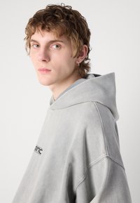 Sudadera gris de gran tamaño hecha de tejido suave, con una capucha grande y un pequeño logo en la parte frontal. Textura suave con sutiles detalles de costura.