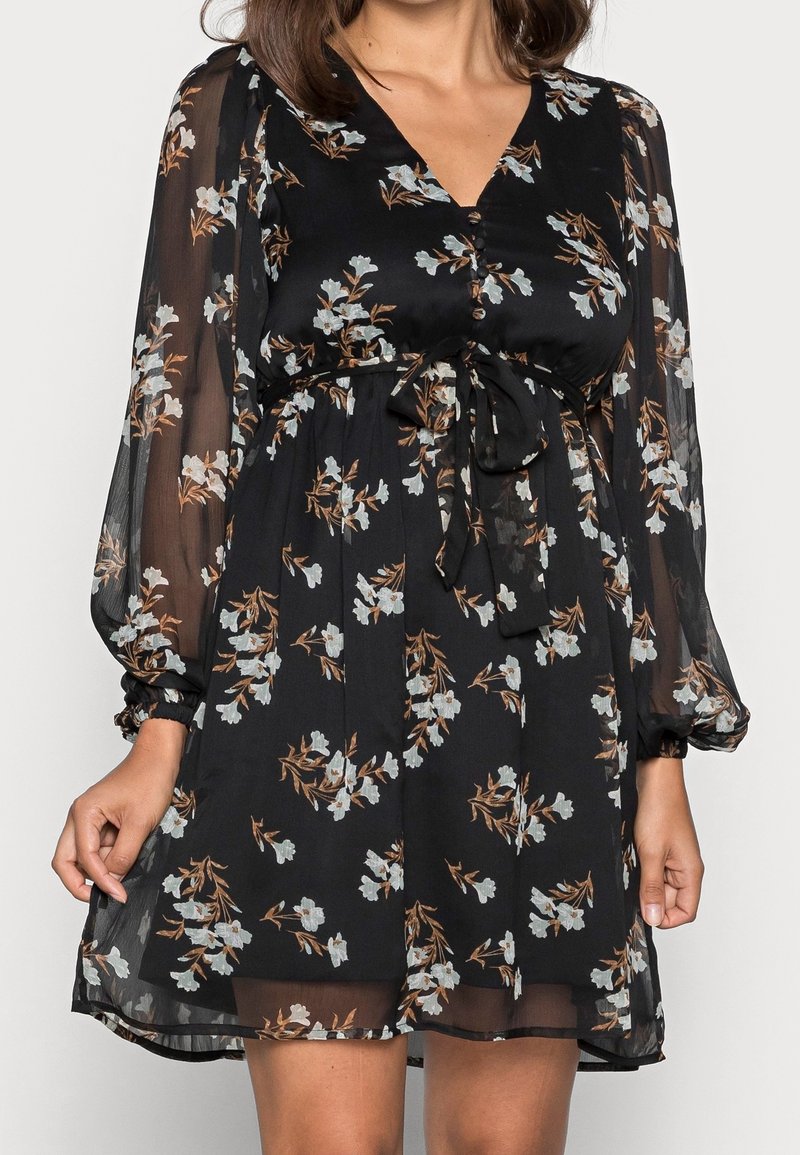 Robe noire fleurie avec manches longues transparentes, col en V à boutons et ceinture nouée à la taille, portée mi-cuisse par une femme.