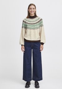 Knit sweater in cream with green and brown geometric patterns, featuring a high neckline and voluminous sleeves, paired with wide-leg denim jeans.