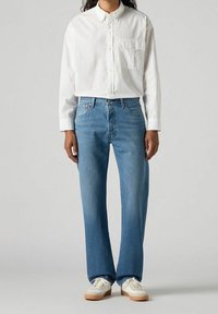 Camisa blanca de botones, jeans azul rectos y zapatillas blancas con suela beige. La tela es suave y los jeans tienen un lavado desteñido.
